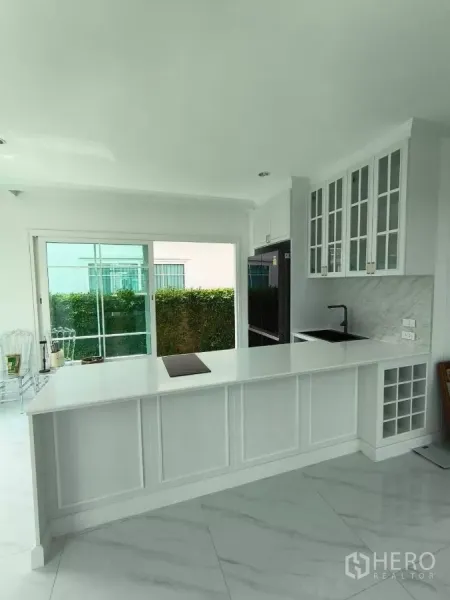 บ้าน ให้เช่าในBang Phli, Samut Prakan - ครัวสีขาวพร้อมเคาน์เตอร์ยาว ตู้กระจก และประตูกระจกบานเลื่อนออกสู่สวน