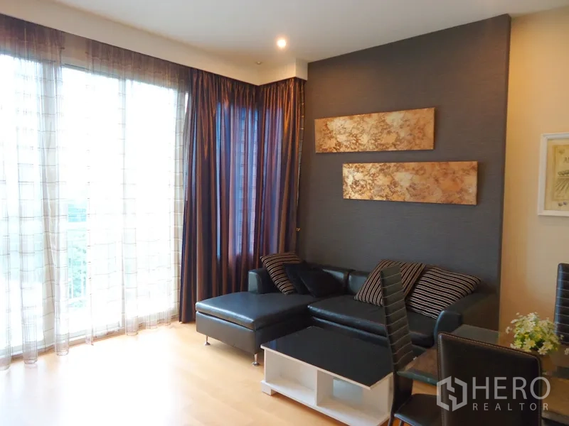 Condominium for rent in Chatuchak, Bangkok - Corner living room featuring an L-shaped leather sofa, floor-to-ceiling windows and dark drapes.
