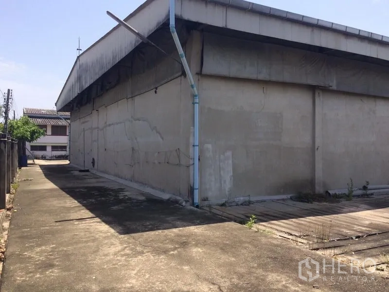 Warehouse for rent in Samut Prakan - Side exterior of the warehouse with narrow driveway and drainage pipe.