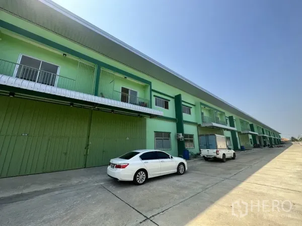 货栈 出租在Pathum Thani - 整排绿色仓库单元，车道宽敞，前方停有车辆。