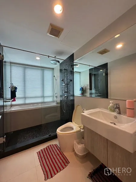Condominium for rent in Khlong Toei, Bangkok - Main bathroom with glass-enclosed shower and bathtub combo, wide mirror and vanity.