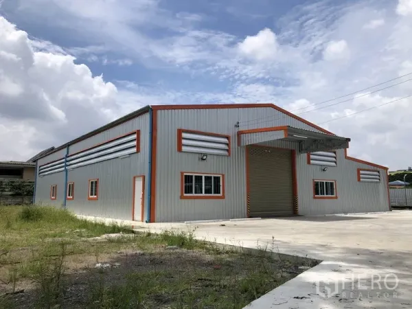 货栈 出租在Mueang Nonthaburi, Nonthaburi - 新建钢结构仓库外观，橙色边框，卷帘门与宽阔混凝土院场，蓝天之下。