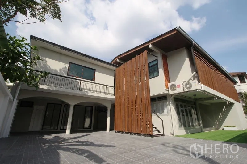 Detached House for rent in Chatuchak, Bangkok - Rear courtyard view of two‑storey home with vertical timber screens, balcony, tiled patio, and small lawn under blue sky.