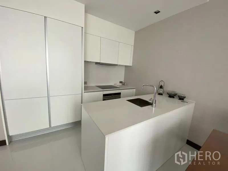 Condominium for rent in Khlong Toei, Bangkok - Minimalist white kitchen with an island sink, built-in oven and ample storage cabinetry.