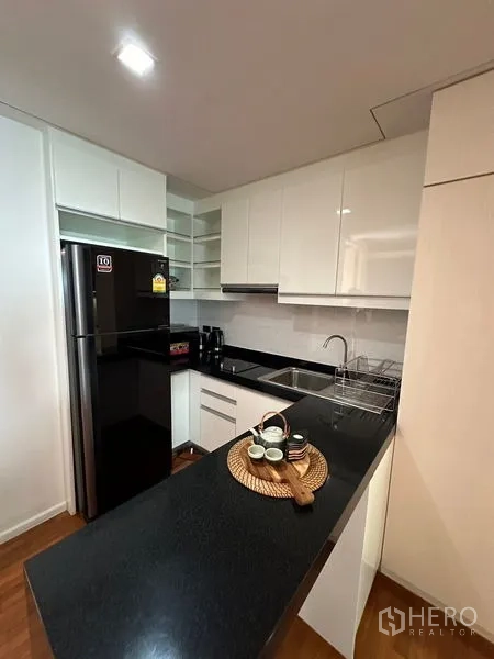 Apartment for rent in Khlong Toei, Bangkok - Compact white kitchen with black stone countertop and large refrigerator.