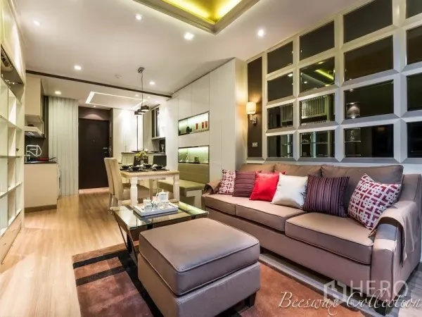 Condominium for rent in Watthana, Bangkok - Contemporary living and dining area with sofa, ottoman, glass coffee table, built‑in shelving and an open kitchen under recessed lighting.