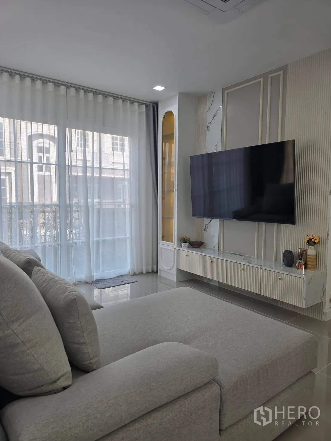 Semi-detached House for rent in Mueang Samut Prakan, Samut Prakan - Bright living room with large sectional sofa, wall-mounted TV and marble media cabinet beside floor-to-ceiling sheer curtains.