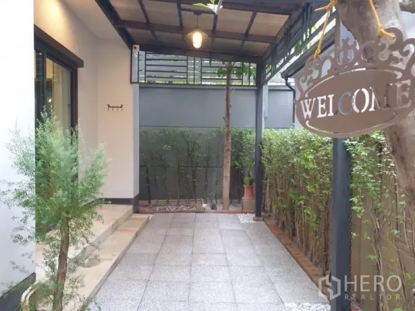 Detached House for rent in Bang Kapi, Bangkok - Shaded patio at the side of the house with tiled floor, hedges, and a hanging welcome sign.