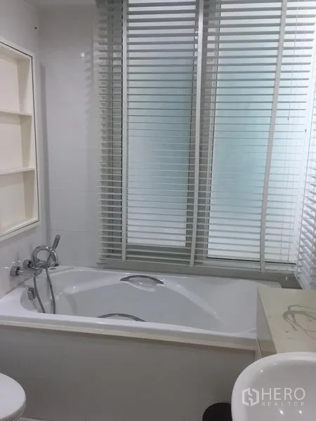 Condominium for rent in Khlong Toei, Bangkok - White bathroom with bathtub, handheld shower, sink, shelving and window blinds.