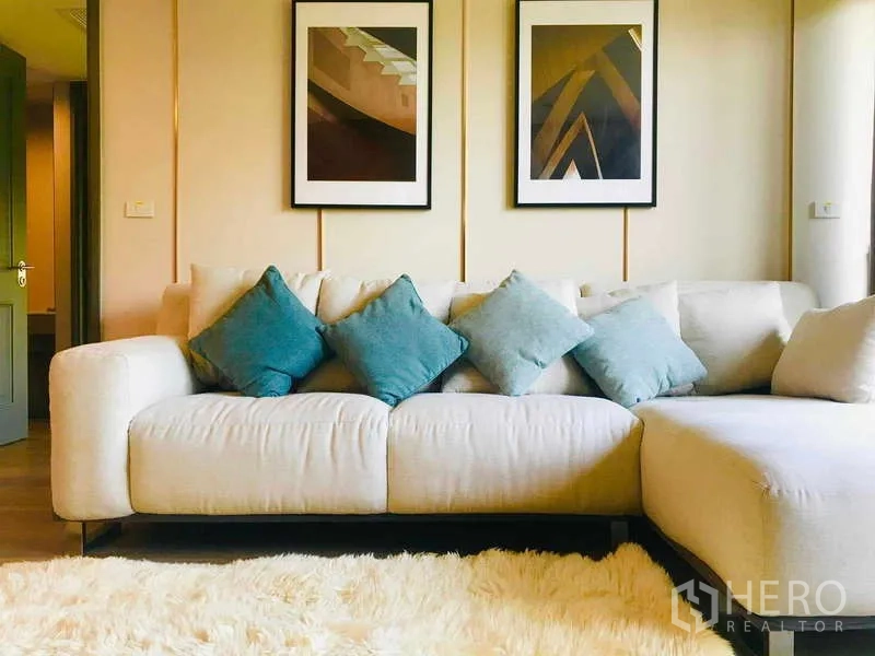 คอนโด ให้เช่า - ห้องนั่งเล่นสว่าง โซฟา L สีครีม หมอนสีฟ้า ภาพแขวน และพรมฟู