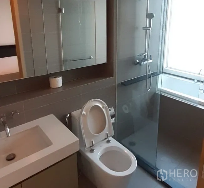 Condominium for rent in Khlong Toei, Bangkok - Stylish gray-tiled bathroom with glass shower enclosure, vanity cabinet, and window for natural light.