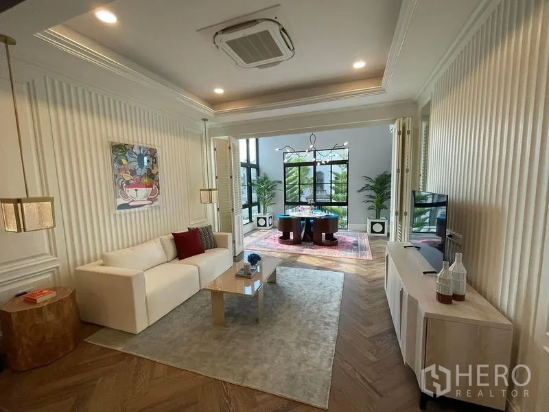 Detached House for rent or sale in Saphan Sung, Bangkok - Family lounge with white sofa, TV console and folding doors opening to a glass-walled sunroom.