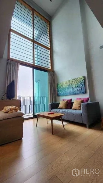 Condominium for rent in Khlong Toei, Bangkok - Loft-style living area with tall windows and sofa by balcony.