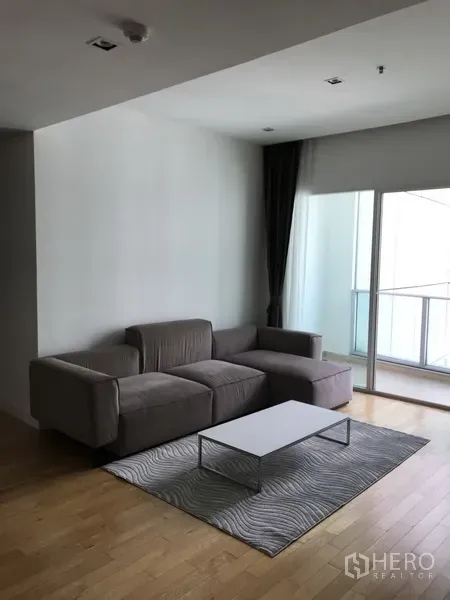 Condominium for rent in Khlong Toei, Bangkok - Living area with gray sectional sofa, low coffee table, and floor-to-ceiling balcony doors.