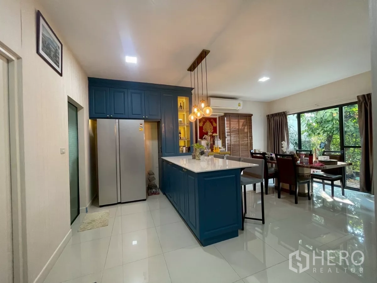 บ้าน ขายในBang Phli, Samut Prakan - ครัวและพื้นที่ทานอาหารแบบเปิด ตู้ครัวสีน้ำเงิน ไอส์แลนด์ และไฟห้อย มองเห็นสวน