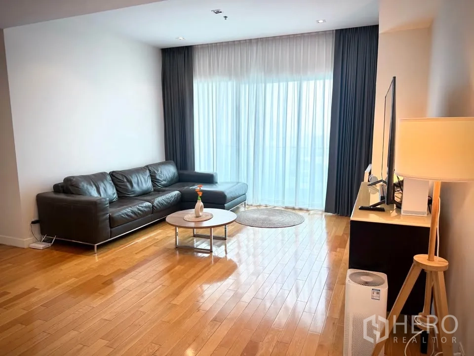 Condominium for rent or sale in Khlong Toei, Bangkok - Living room with black leather sectional sofa, TV console, and floor-to-ceiling window.