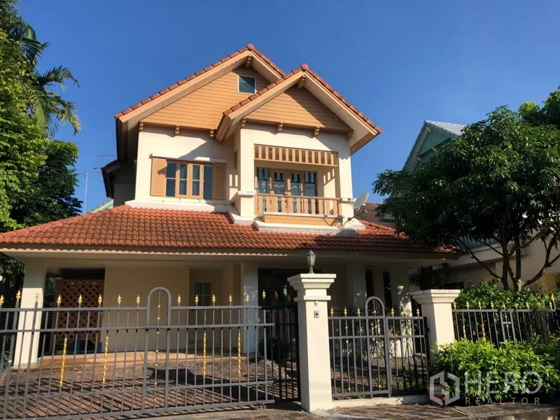 Detached House for rent in Saphan Sung, Bangkok - Front view of a two-storey detached house with red tile roof, balcony and gated entrance surrounded by greenery.