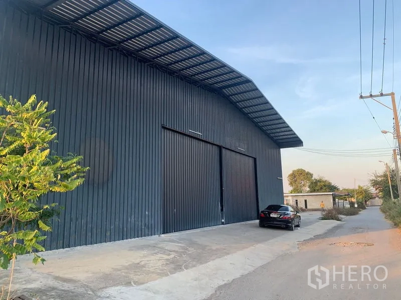 Warehouse for rent in Khan Na Yao, Bangkok - Front exterior of metal-clad warehouse with large sliding doors, covered canopy, driveway, and quiet lane.