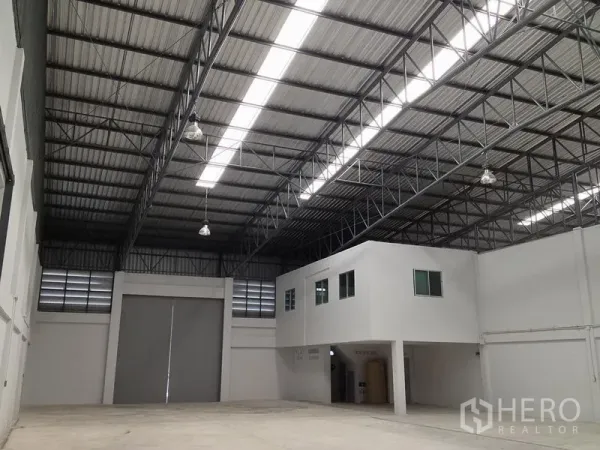 Warehouse for rent in Si Racha, Chon Buri - High-ceiling warehouse interior with steel trusses, skylight strips, a built-in two-storey office and a large roller door.