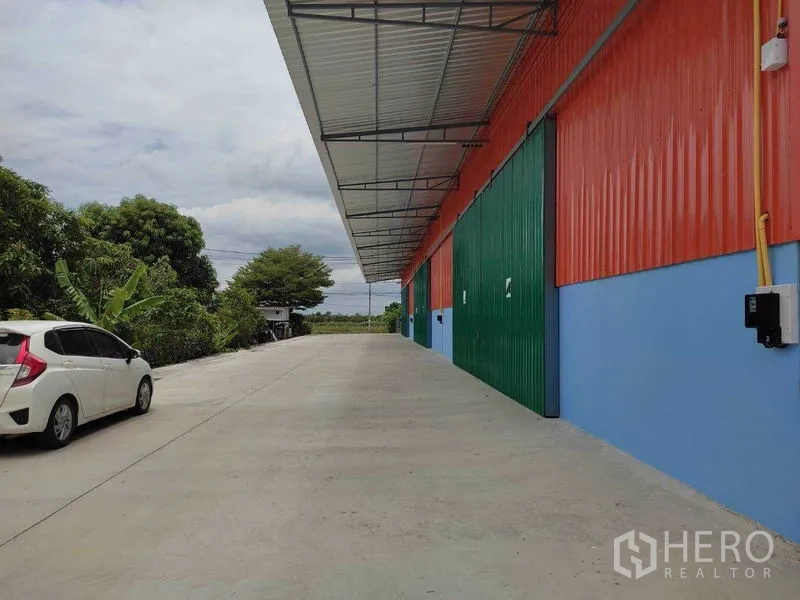 Warehouse for rent in Bang Yai, Nonthaburi - Wide concrete driveway beside a red-and-blue warehouse with green sliding doors and a covered loading canopy.
