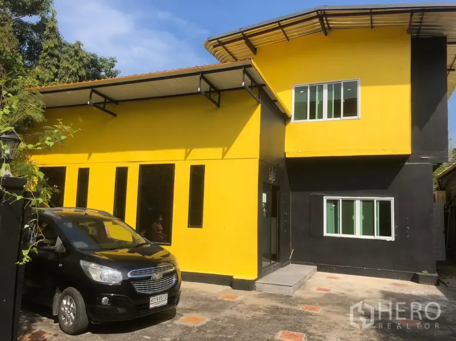 Detached House for rent in Khlong Toei, Bangkok - Two-storey yellow-and-black detached house with sliding windows, paved driveway and on-site parking under a clear sky.