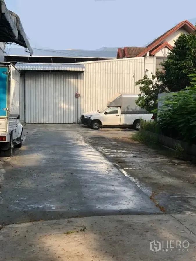 Warehouse for rent in Wang Thonglang, Bangkok - Concrete driveway leading to a metal-clad warehouse with a large sliding door and parked delivery vehicles.