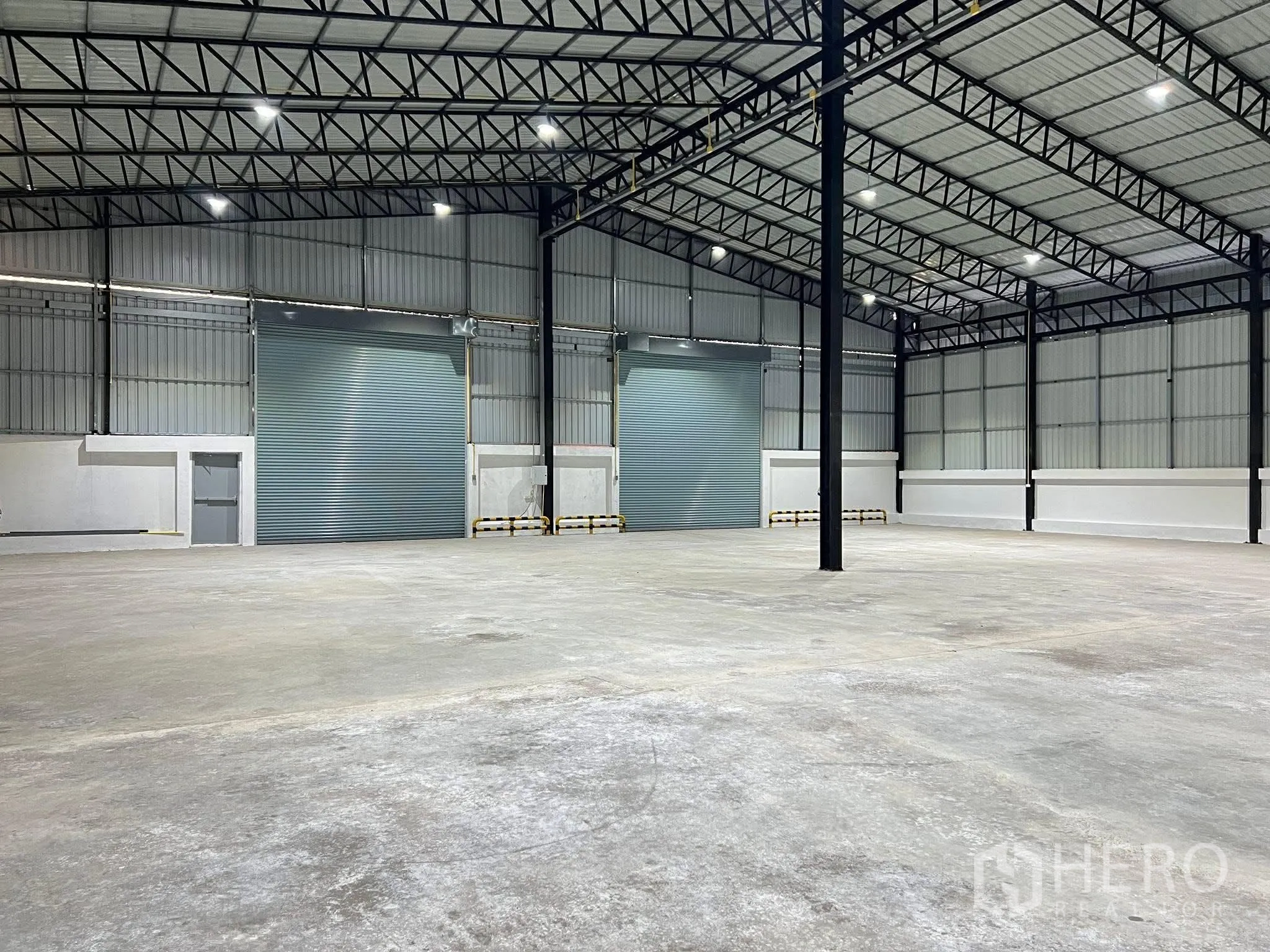 Warehouse for rent in Nong Chok, Bangkok - Open warehouse interior with high steel truss roof, concrete floor, and two large roller shutters.