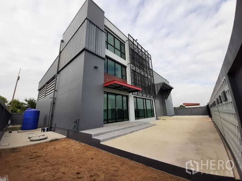 Factory for rent or sale in Lam Luk Ka, Pathum Thani - Side angle of the property with wide concrete driveway, gated entrance and loading bay.