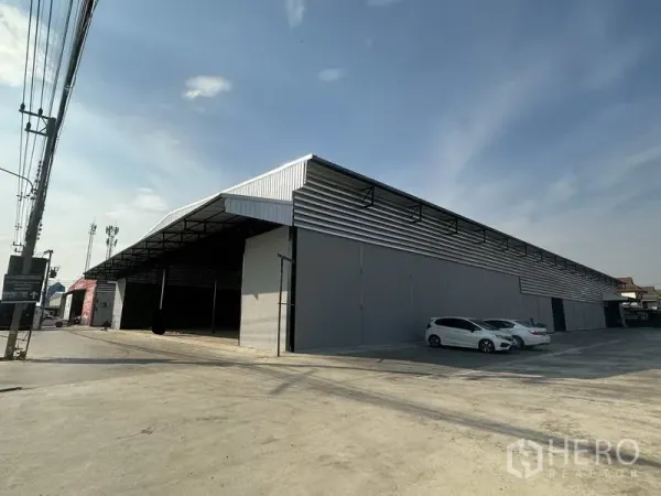 Warehouse for rent in Lam Luk Ka, Pathum Thani - Angled exterior of a long grey warehouse featuring a covered loading area and cars parked on the paved yard.
