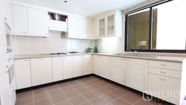Apartment for rent in Khlong Toei, Bangkok - Bright U-shaped kitchen with white cabinets, cooktop, ample counter space and large window.