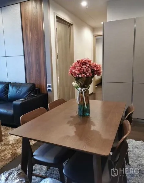 Condominium for rent or sale in Watthana, Bangkok - Dining area with wooden table, four chairs and floral centerpiece next to living zone.