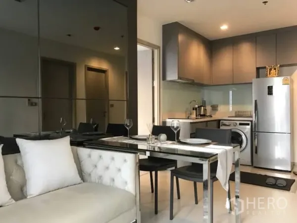 Condominium for rent or sale in Khlong Toei, Bangkok - Open-plan living and dining area beside a modern kitchenette with a mirrored wall.