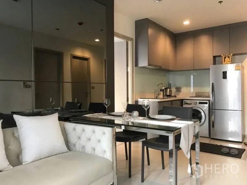 Condominium for rent or sale in Khlong Toei, Bangkok - Open-plan living and dining area beside a modern kitchenette with a mirrored wall.