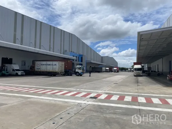 货栈 出租在Chachoengsao - 仓库之间的大型混凝土货车场地，集装箱卡车在雨棚下作业。