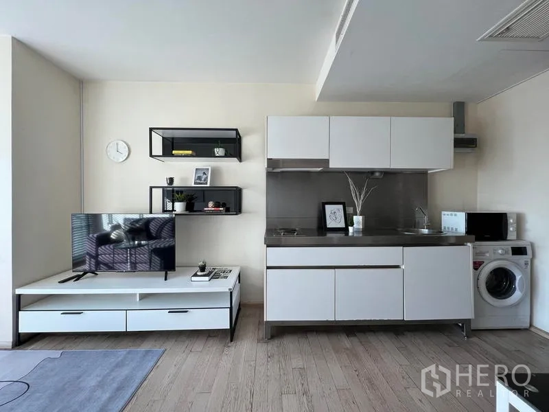 Condominium for rent in Khlong Toei, Bangkok - Open-plan living room with TV unit and compact white kitchen with washer.