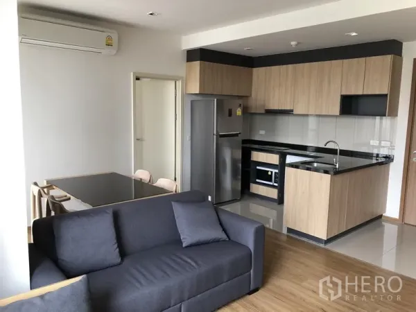 Condominium for rent in Watthana, Bangkok - Open‑plan living area with a grey sofa, dining table, and a wood‑finished kitchen with black countertops and built‑in appliances.
