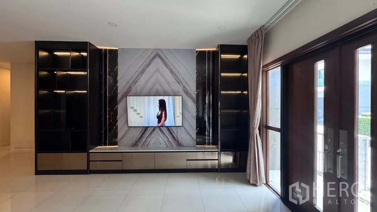 Detached House for rent or sale in Saphan Sung, Bangkok - Living room with marble feature wall, built‑in black shelving and a wall‑mounted TV beside large wooden entry doors and curtains.