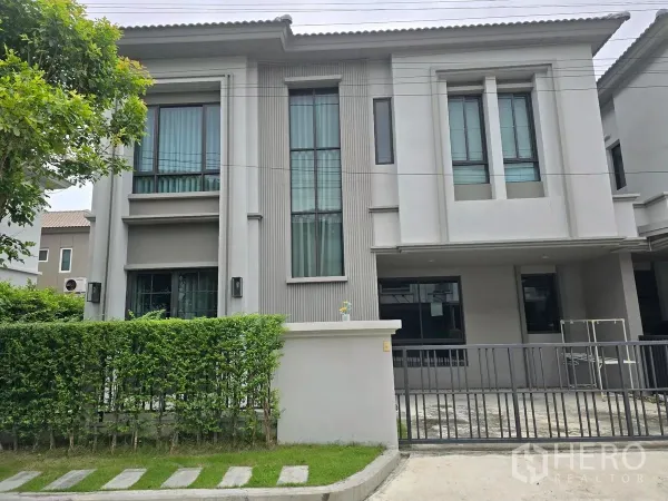 Detached House for rent in Khlong Luang, Pathum Thani - Modern two-storey detached house exterior with tall windows, green hedge, and sliding gate in Khlong Luang, Pathum Thani.
