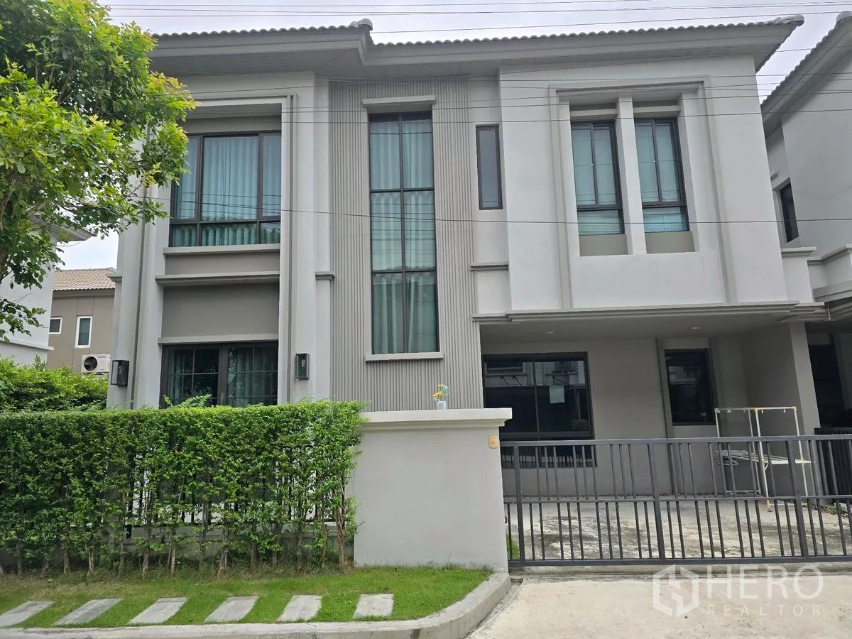 Detached House for rent in Khlong Luang, Pathum Thani - Modern two-storey detached house exterior with tall windows, green hedge, and sliding gate in Khlong Luang, Pathum Thani.