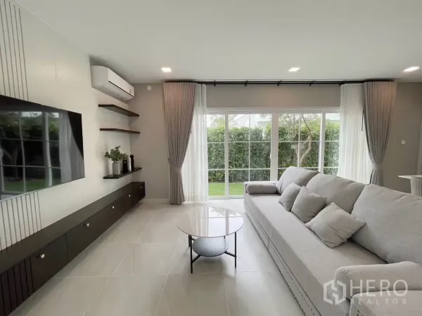 Detached House for rent in Bang Phli, Samut Prakan - Bright living room with large gray sofa, TV wall, and sliding glass doors opening to the garden.