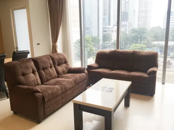 公寓 出租在Bang Rak, Bangkok - 采光充足的客厅，落地窗旁两张棕色沙发与咖啡桌，眺望城市景观。