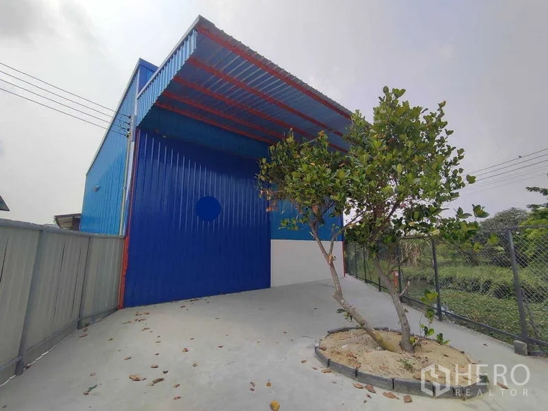 货栈 出租在Pak Kret, Nonthaburi - 仓库前车道与带雨棚的装卸区，蓝色金属外墙，侧面围栏。