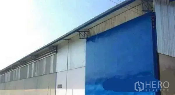 Factory for rent in Mueang Nakhon Pathom, Nakhon Pathom - Factory side exterior showing a large blue metal sliding door and sheet‑metal roof.
