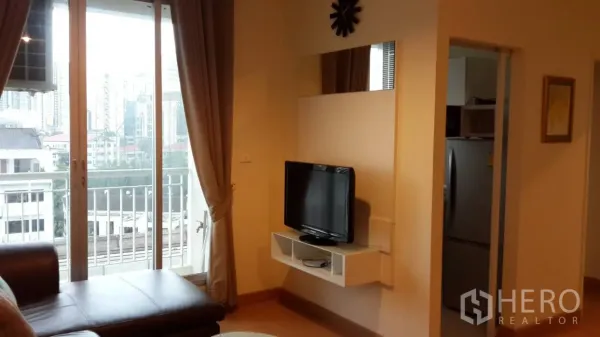 Condominium for rent in Watthana, Bangkok - Bright living room with sliding glass doors to a balcony, city view, curtains, and a TV on a floating shelf.
