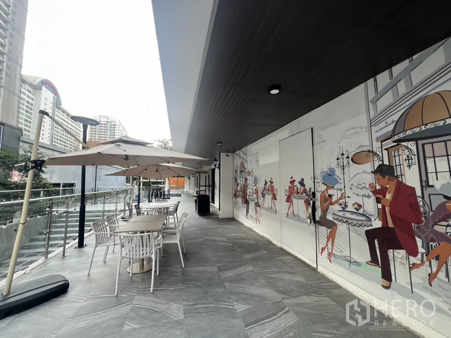 Retail Space for rent - Promenade with café-style mural, outdoor tables and umbrellas along a glass-railed walkway.