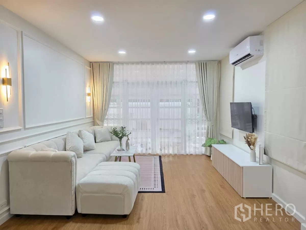 Detached House for rent in Huai Khwang, Bangkok - Modern living area with sectional sofa, wall-mounted TV, air-conditioning and floor-to-ceiling drapes.