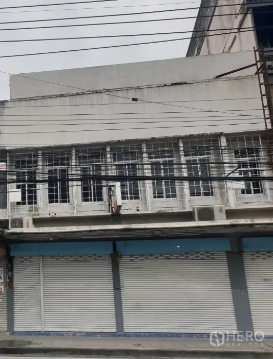 อาคารพาณิชย์ ให้เช่าในWatthana, Bangkok - ด้านหน้าอาคารพาณิชย์ติดถนนในกรุงเทพฯ ประตูม้วนโลหะปิด หน้าต่างชั้นบนแบบกรงเหล็กและสายไฟเหนือถนน.