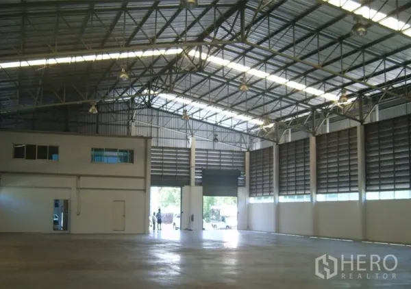 Warehouse for rent in Samut Prakan - Spacious high-clearance warehouse interior with steel trusses, skylights, and a large roller door.