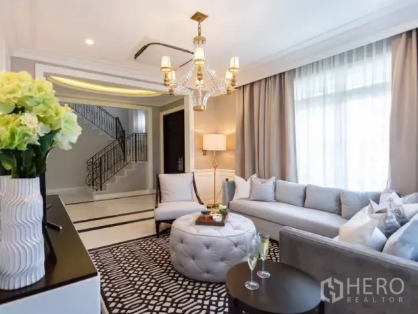 Detached House for rent in Mueang Samut Prakan, Samut Prakan - Cozy living room with L‑shaped sofa, chandelier and view to the staircase entry hall.