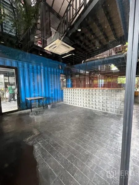 零售空间 出租 - 透过玻璃看到的开放式店铺，高挑空间、蓝色集装箱墙与灯光吧台。